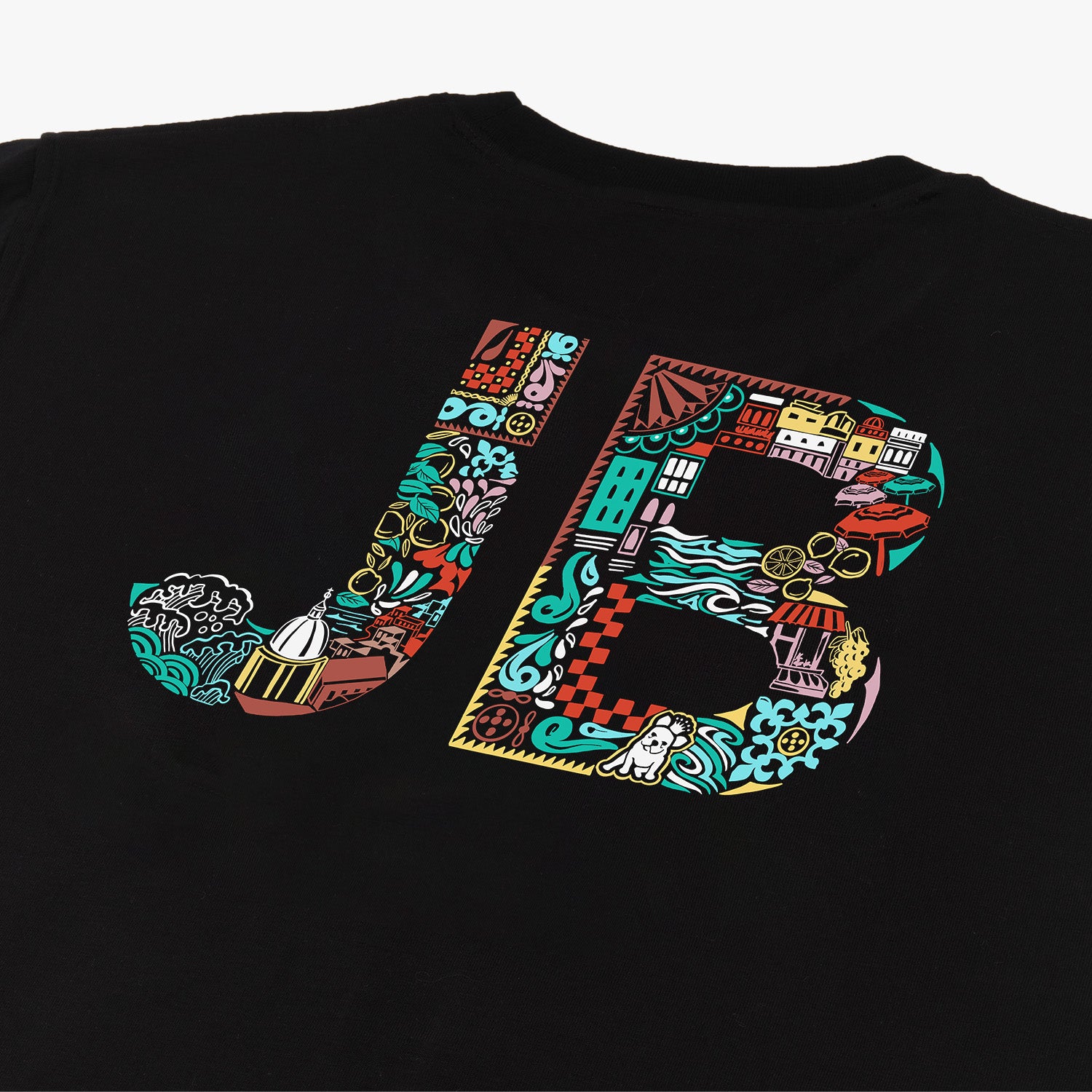 Close-up of the back of a black t-shirt featuring large, colorful 'JB' initials. The letters are filled with intricate illustrations including buildings, waves, gears, and various abstract designs. The vibrant colors and detailed patterns create a bold, eye-catching design.