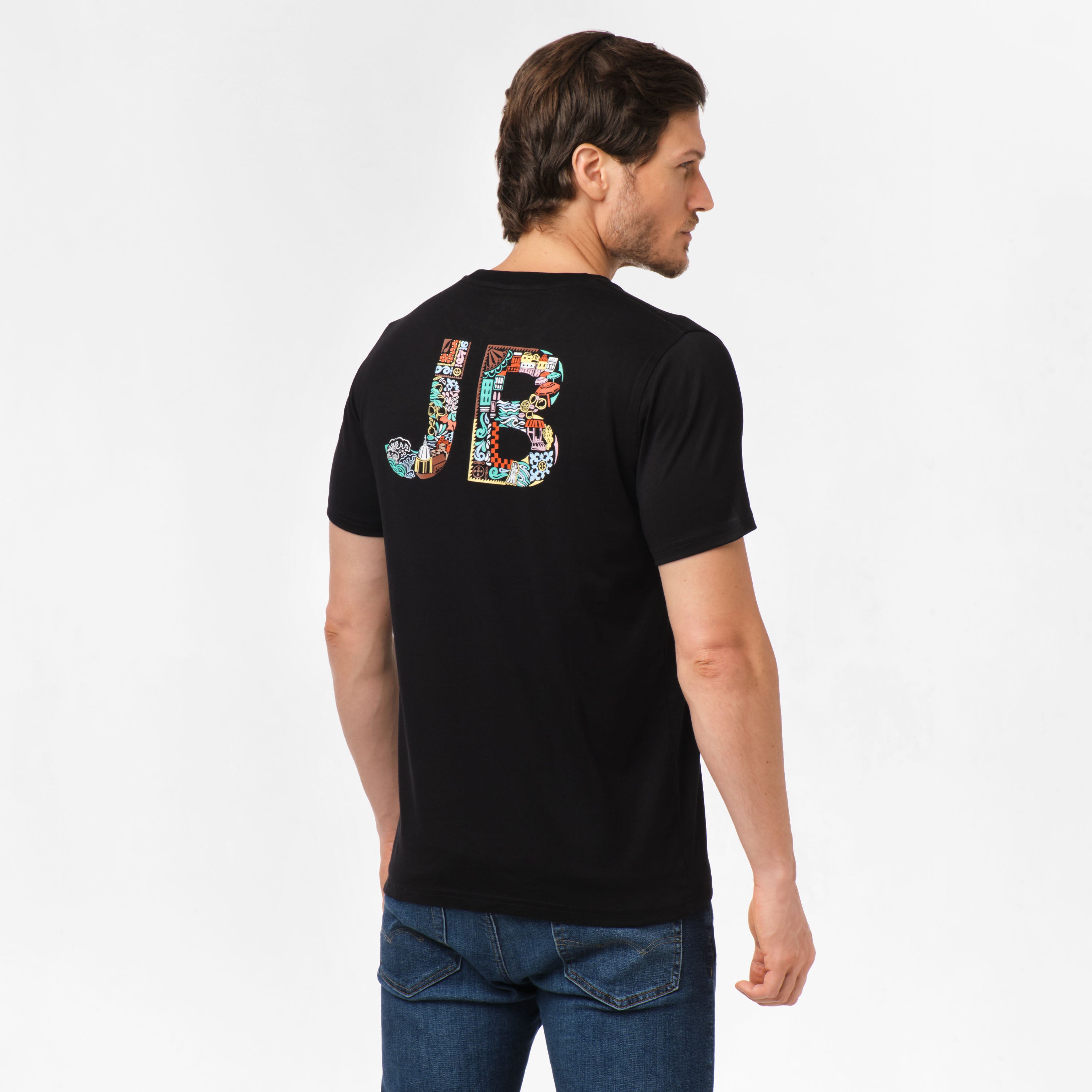 Man wearing a black t-shirt with large, multicolored 'JB' letters on the back. The letters are composed of various patterns and designs. The man is shown from the back, standing with his head turned to the side, wearing blue jeans.