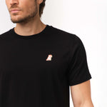 Close-up view of a black t-shirt featuring a small embroidered dog logo on the left chest. The logo is orange and white, adding a subtle design element to the plain black t-shirt.