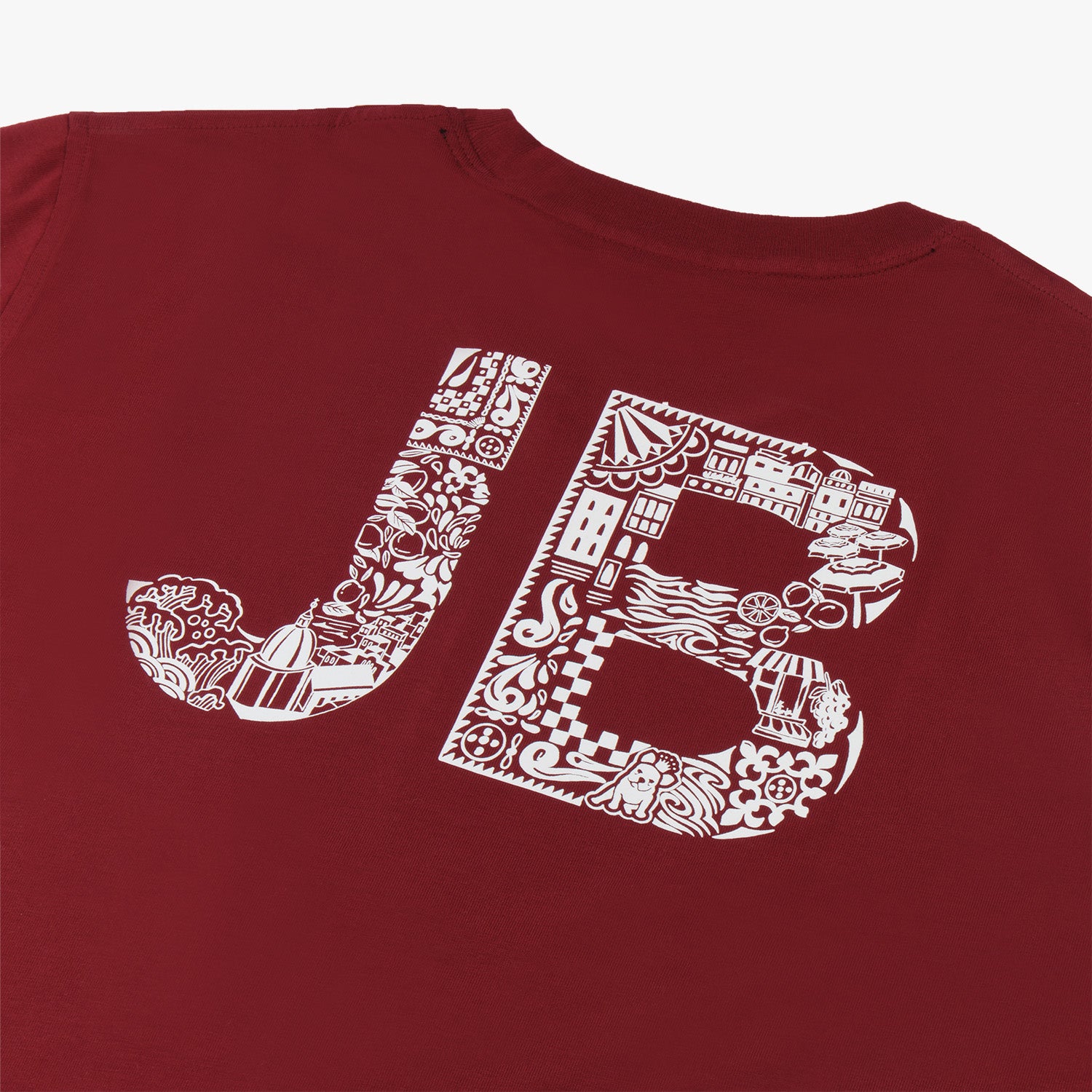 Close-up of the back design of a maroon t-shirt featuring large white 'JB' initials. The initials are intricately filled with detailed illustrations, including elements like buildings, plants, and various decorative patterns, creating a vibrant and artistic graphic. The design combines urban and nature-inspired motifs within the letters, giving the shirt a distinctive and creative flair.