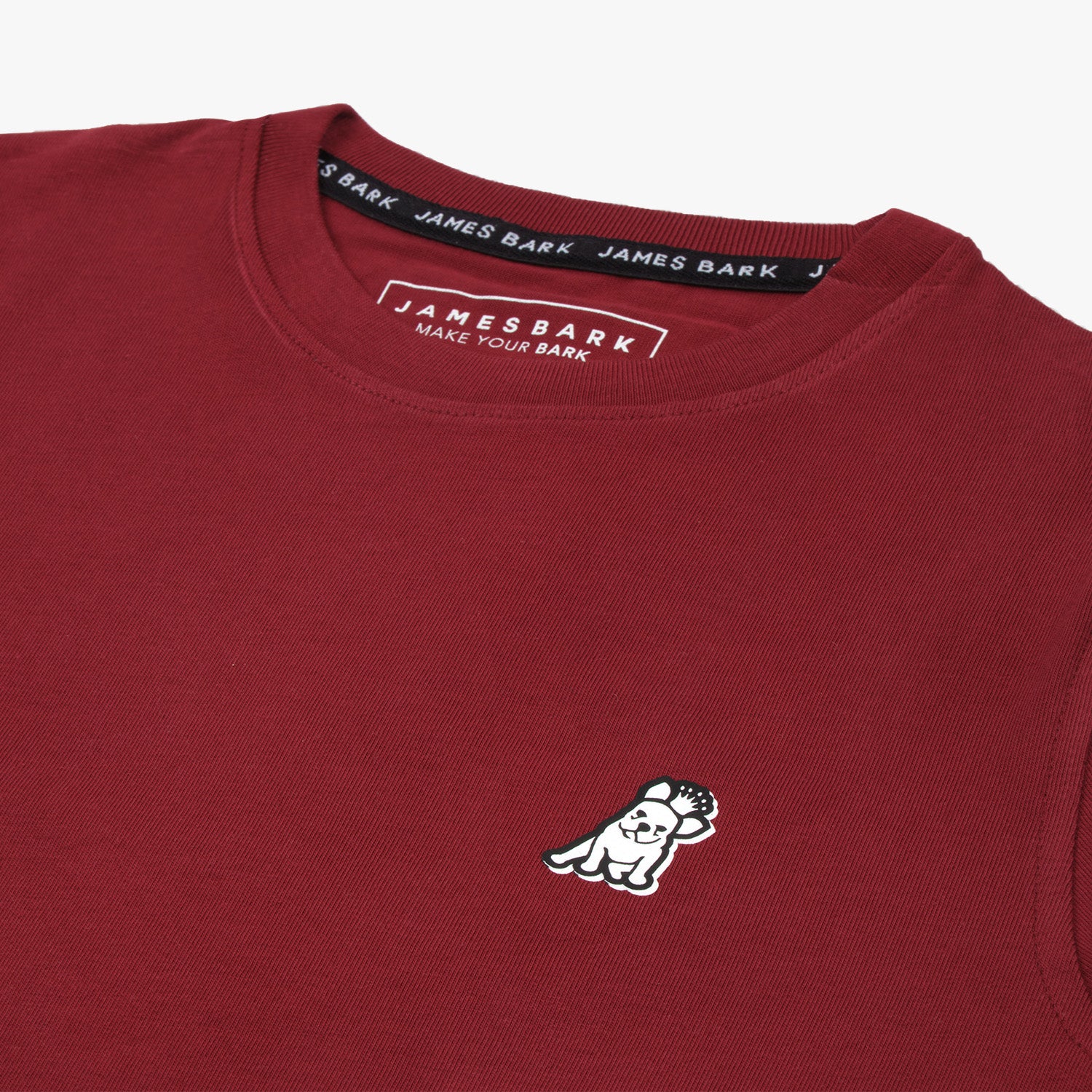 Detailed close-up of a maroon t-shirt featuring a small embroidered patch of a white dog raising one paw on the left chest area. The collar tag reads 'JAMES BARK - MAKE YOUR BARK' in white letters, and the inner collar lining is branded with 'JAMES BARK' text. The fabric has a clean and minimalist design.