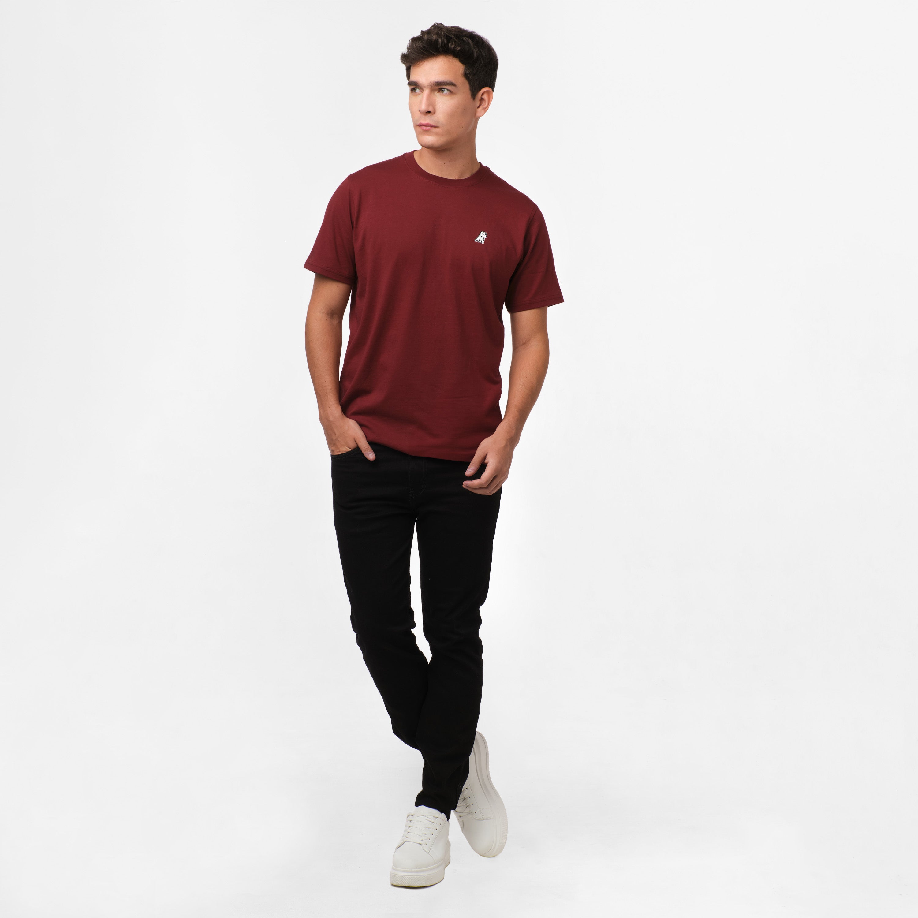 A man wearing a casual maroon t-shirt with a small logo on the chest, paired with black pants and white sneakers. The look is relaxed yet stylish, with the model standing confidently, hands in pockets, and slightly looking to the side. The outfit is simple and versatile, suitable for casual outings.