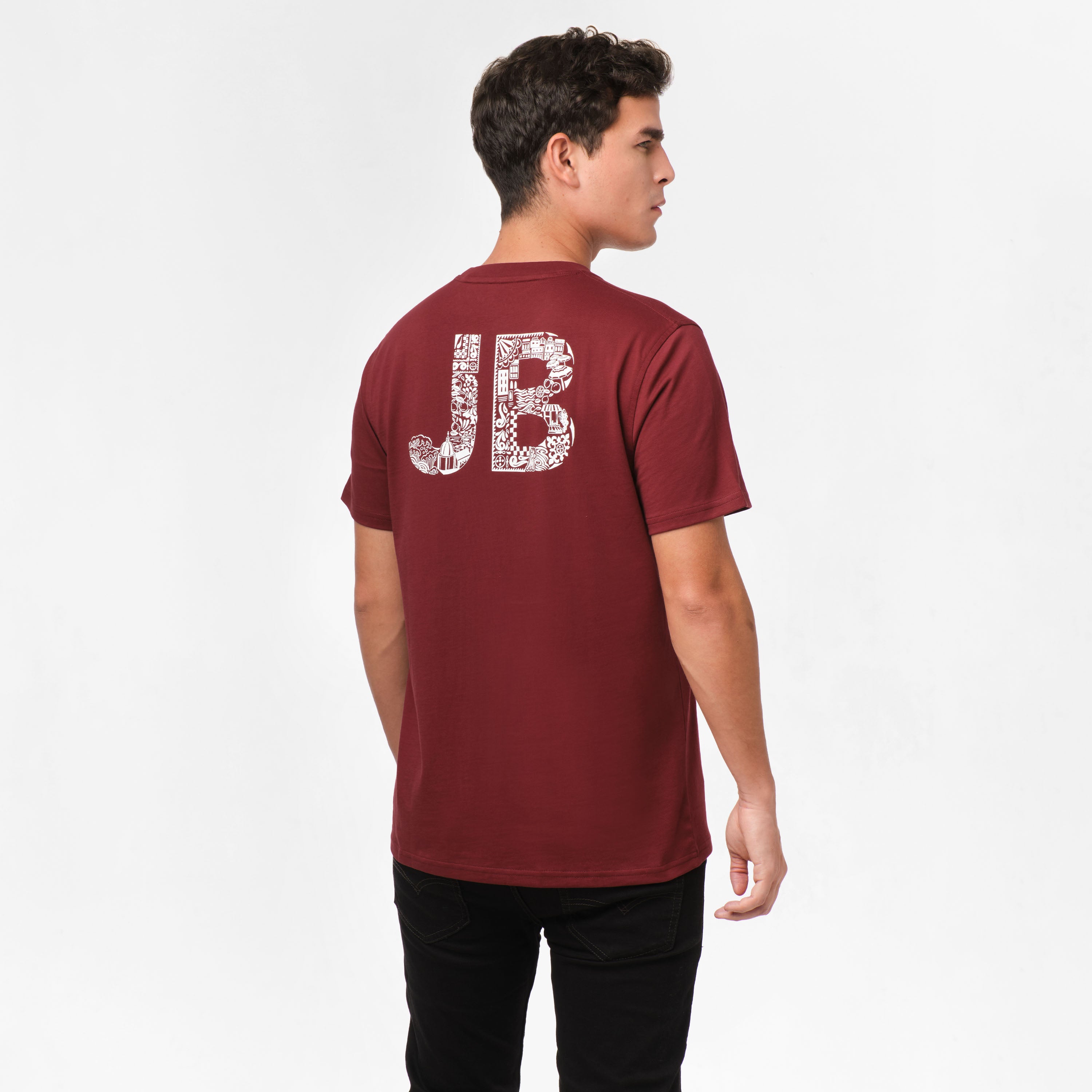 Back view of a man wearing a maroon t-shirt with a large 'JB' graphic on the back. The design is filled with intricate patterns, adding a creative touch to the simple t-shirt. The man is also wearing black pants and standing in a casual pose, facing away from the camera.