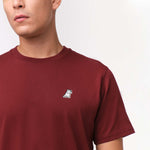 Close-up front view of a maroon t-shirt featuring a small embroidered patch of a white dog with one paw raised, located on the upper left chest area. The shirt has a clean, minimalist design with a relaxed fit.