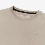 A close-up of a beige T-shirt, highlighting the tonal rubberized logo patch on the upper left chest that reads 'PRIVÉ SOCIETY JAMES BARK EST. 2015.' The neckline features a black inner lining with repeated 'JAMES BARK' branding, and the inside label displays 'JAMES BARK MAKE YOUR BARK' along with material details