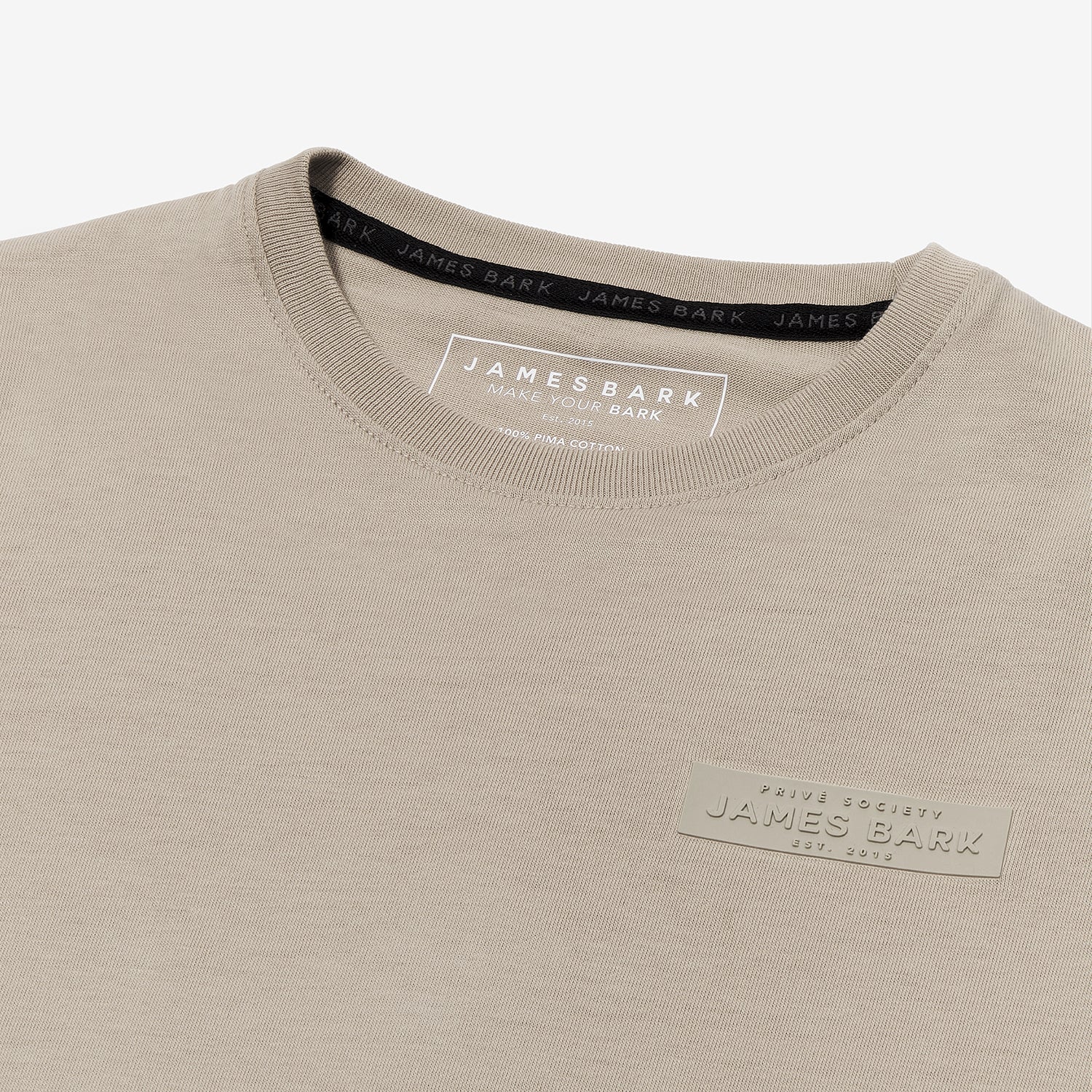 A close-up of a beige T-shirt, highlighting the tonal rubberized logo patch on the upper left chest that reads 'PRIVÉ SOCIETY JAMES BARK EST. 2015.' The neckline features a black inner lining with repeated 'JAMES BARK' branding, and the inside label displays 'JAMES BARK MAKE YOUR BARK' along with material details