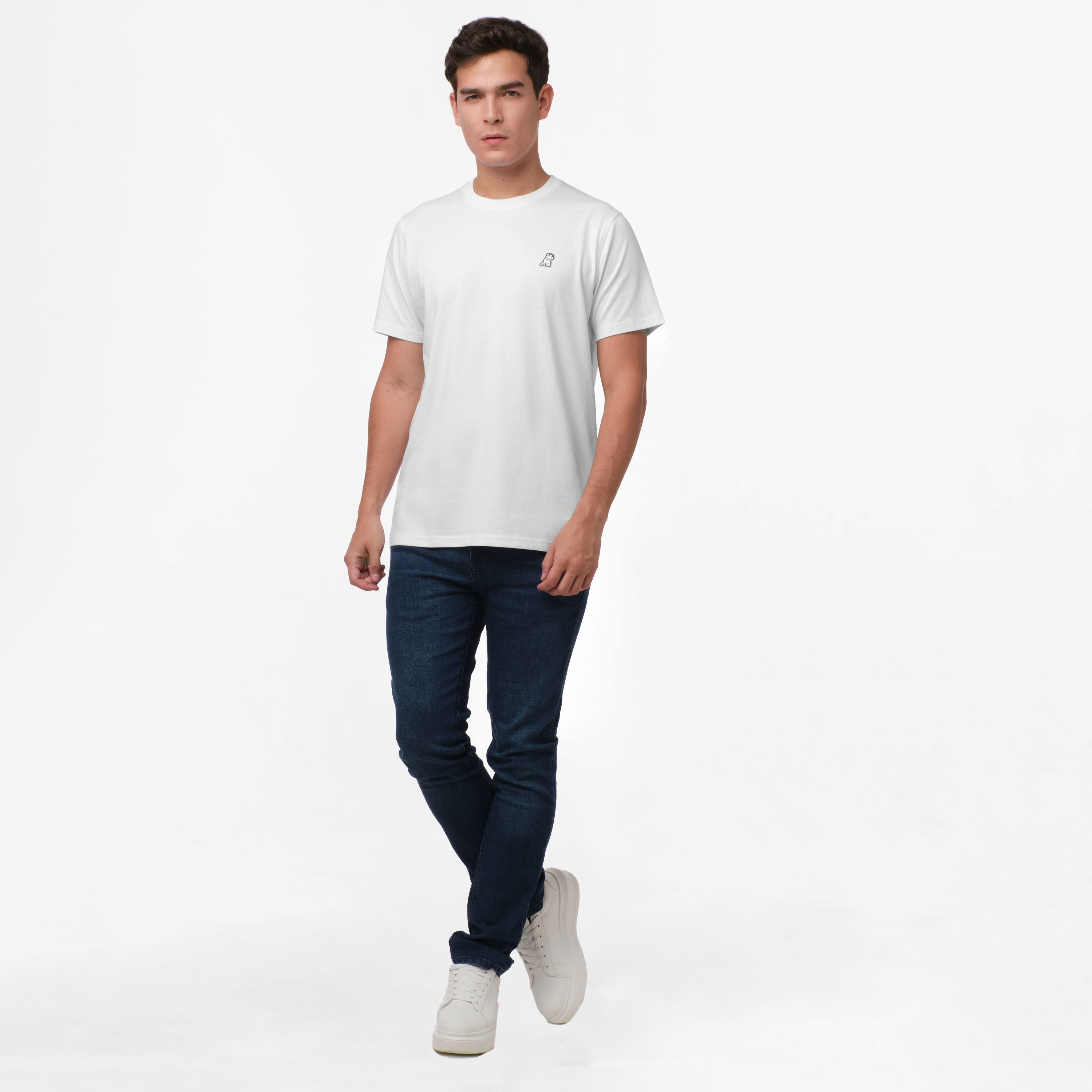 Full-body image of a male model wearing a white t-shirt with a small logo on the chest, paired with dark blue jeans and white sneakers. The model stands in a relaxed pose with one hand in his pocket, exuding a casual and clean look.