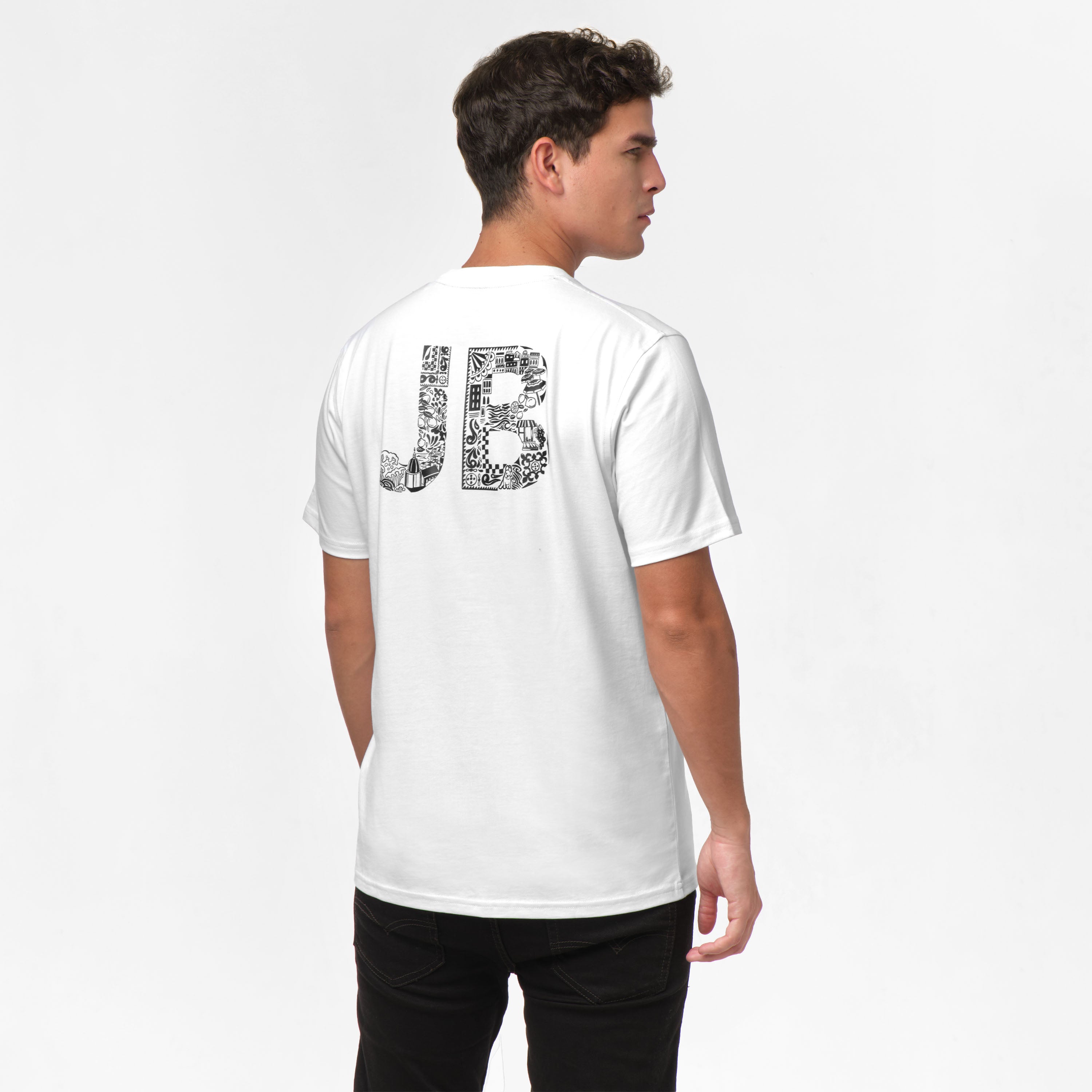 A young man wearing a white t-shirt with a large 'JB' graphic on the back, styled in a detailed and artistic design. He pairs the shirt with black pants, presenting a clean and minimalistic outfit. The model is turned slightly to the side, showcasing the back of the shirt while standing in a relaxed pose.