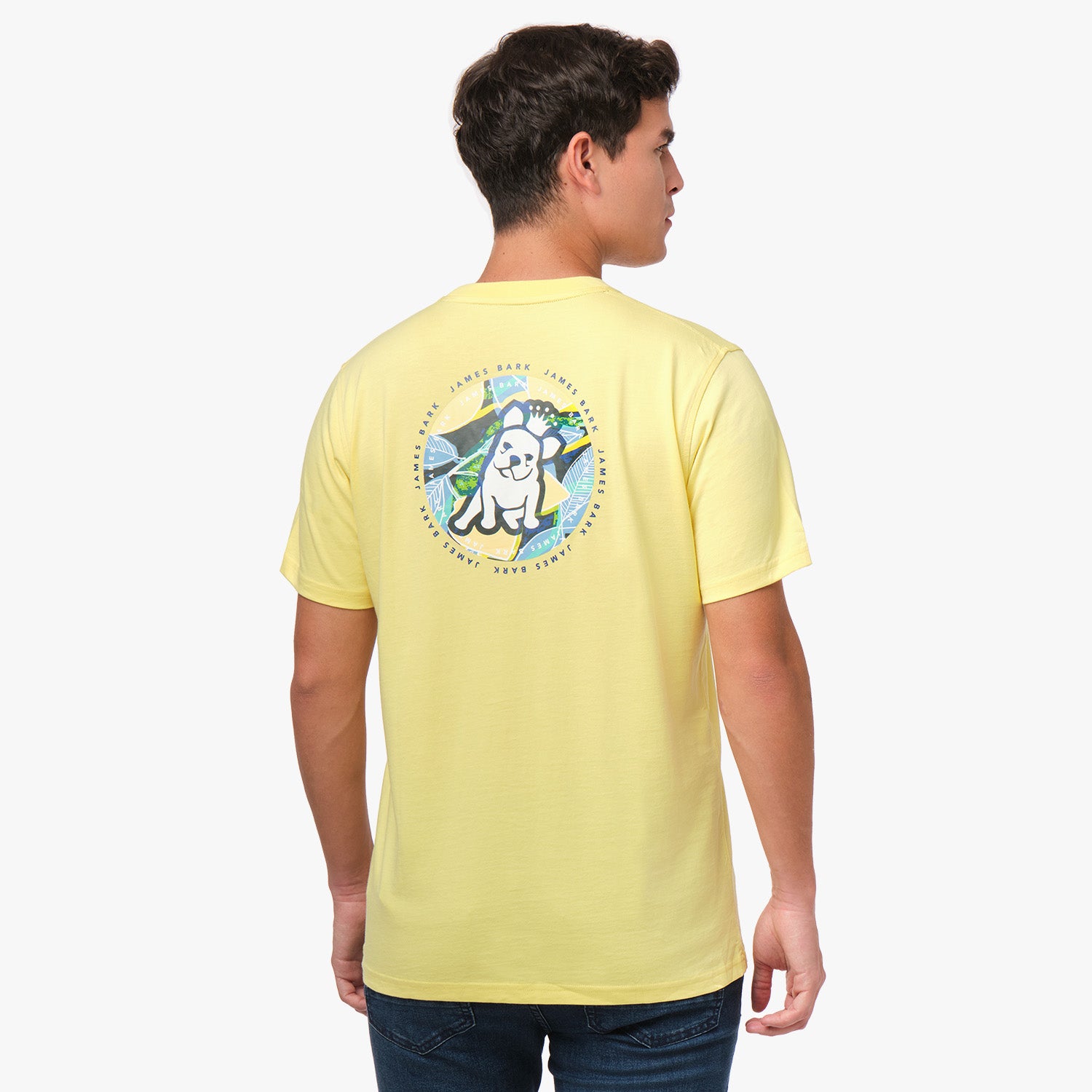 A man is shown from behind, wearing a yellow T-shirt with a large circular graphic on the back. The design features a dog sitting in front of a tropical background with the text "James Bark Privé Society" around the circle. He pairs the shirt with dark pants.