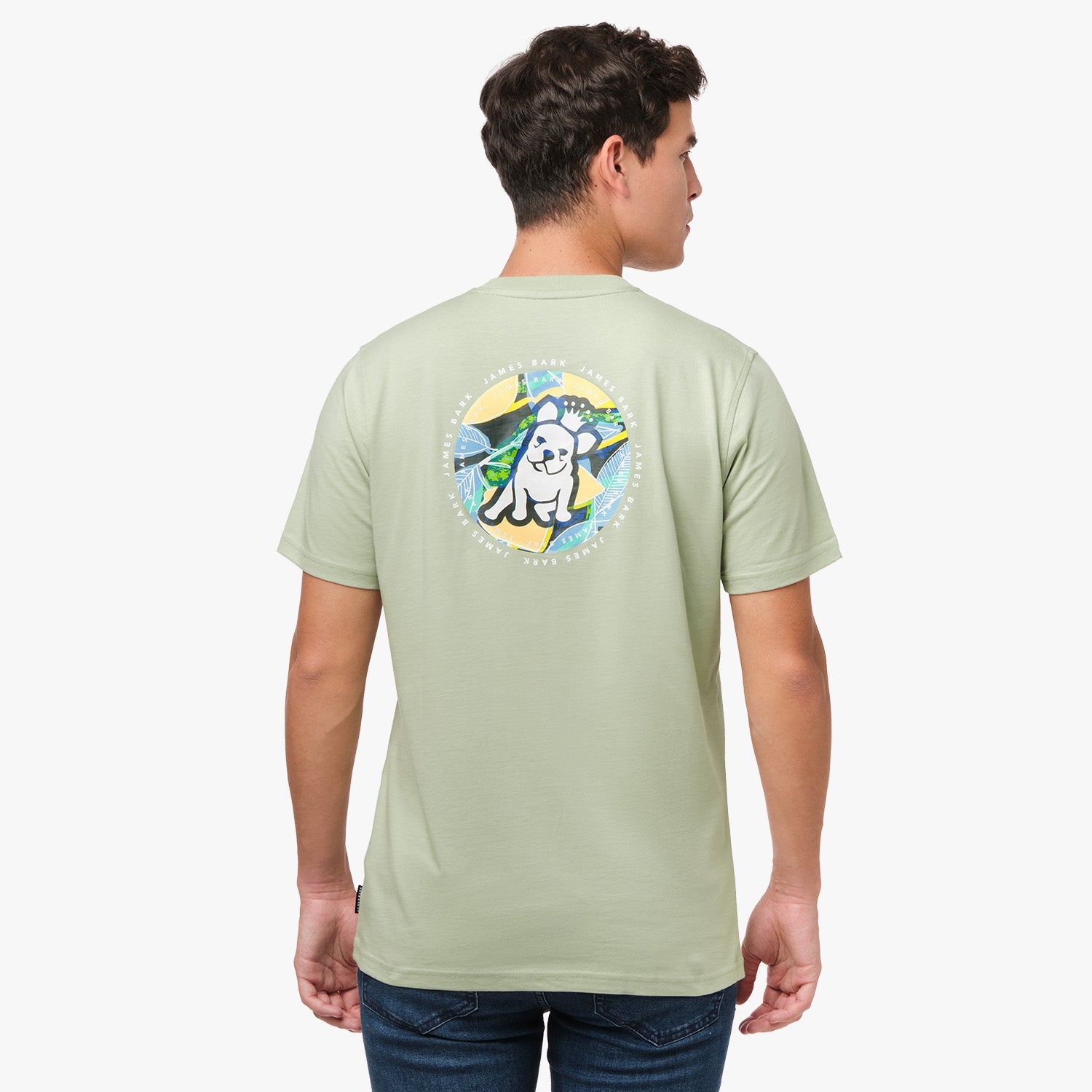 A man is shown from behind, wearing a light green T-shirt with a large circular graphic on the back. The design features a dog sitting in front of a tropical background with the text "James Bark Privé Society" around the circle. He pairs the shirt with dark pants.