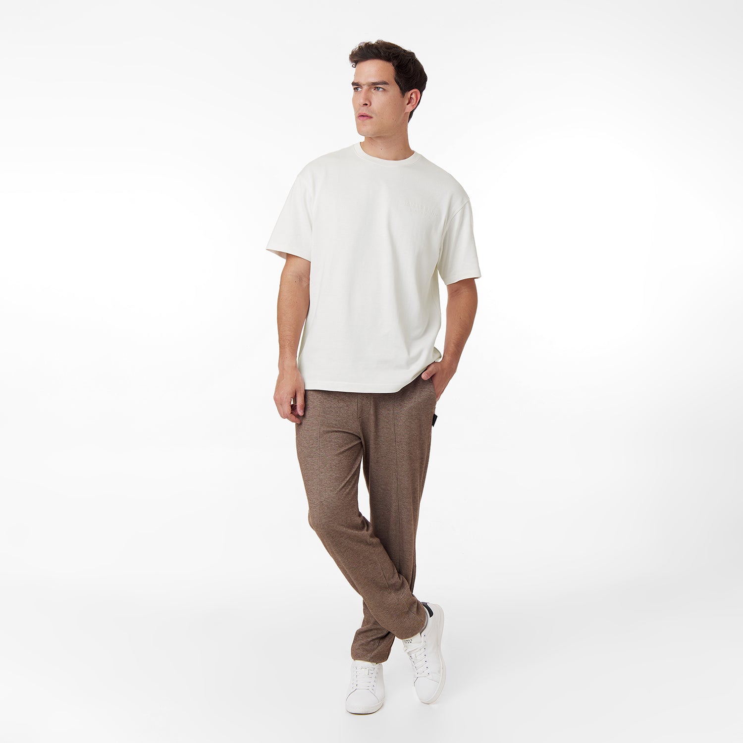 Full-body shot of a male model wearing an oversized off-white t-shirt with an embossed 'JAMES BARK ELEMENTS' logo on the chest. Paired with brown casual pants and white sneakers. Minimalist white background