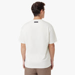 Back view of a male model wearing an oversized off-white t-shirt. Features a minimalist design with a black rectangular 'JAMES BARK' label near the neckline. Paired with brown casual pants. White background