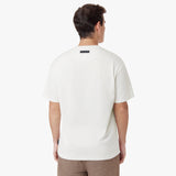 Back view of a male model wearing an oversized off-white t-shirt. Features a minimalist design with a black rectangular 'JAMES BARK' label near the neckline. Paired with brown casual pants. White background