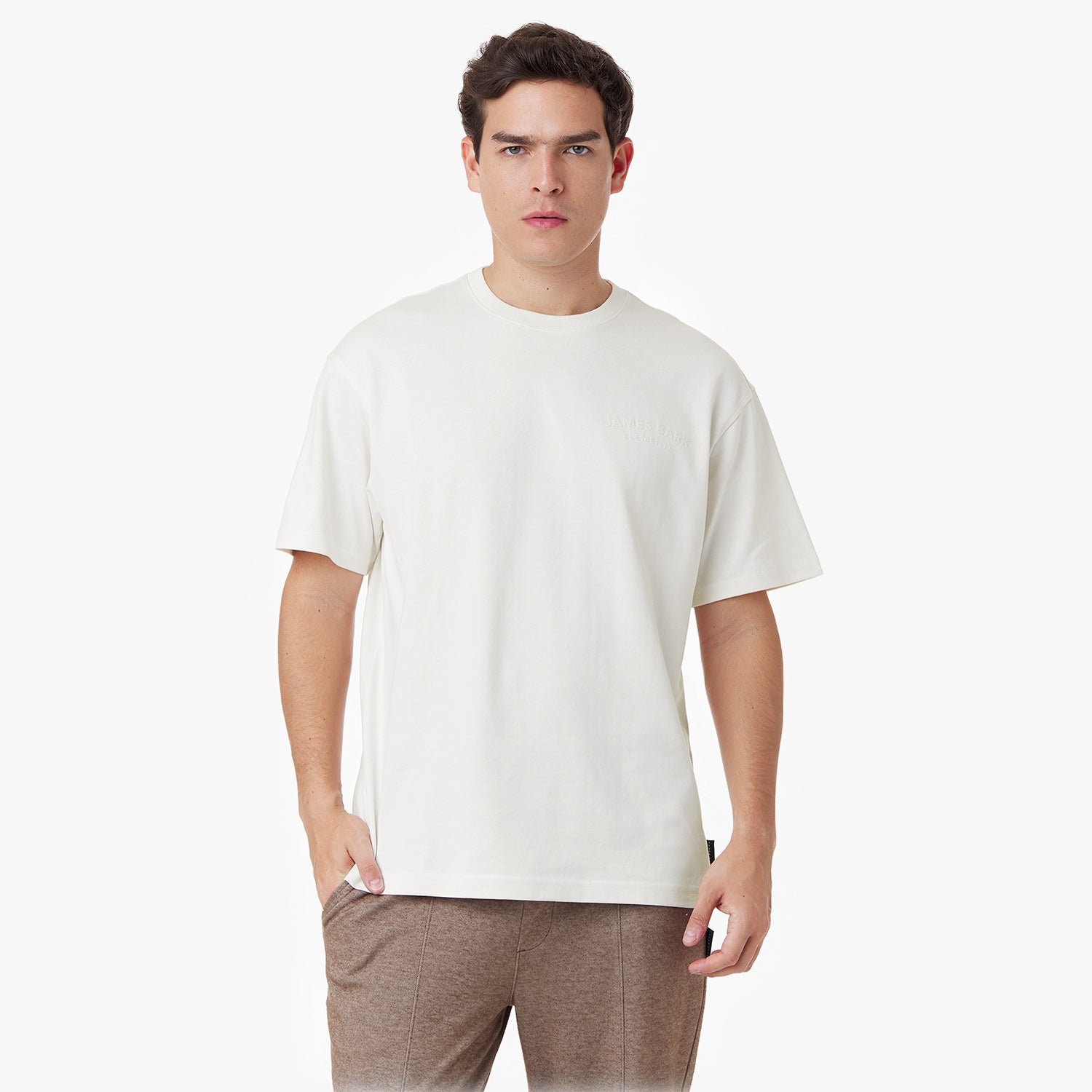 Male model wearing an oversized off-white t-shirt with a minimalist design. Features an embossed 'JAMES BARK ELEMENTS' logo on the chest, a ribbed crew neckline, and short sleeves. Paired with brown casual pants. White background