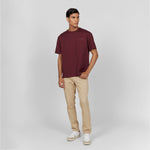 Full-body shot of a male model wearing an oversized burgundy t-shirt with an embossed 'JAMES BARK ELEMENTS' logo on the chest. Paired with beige pants and white sneakers with beige details. Neutral gray background