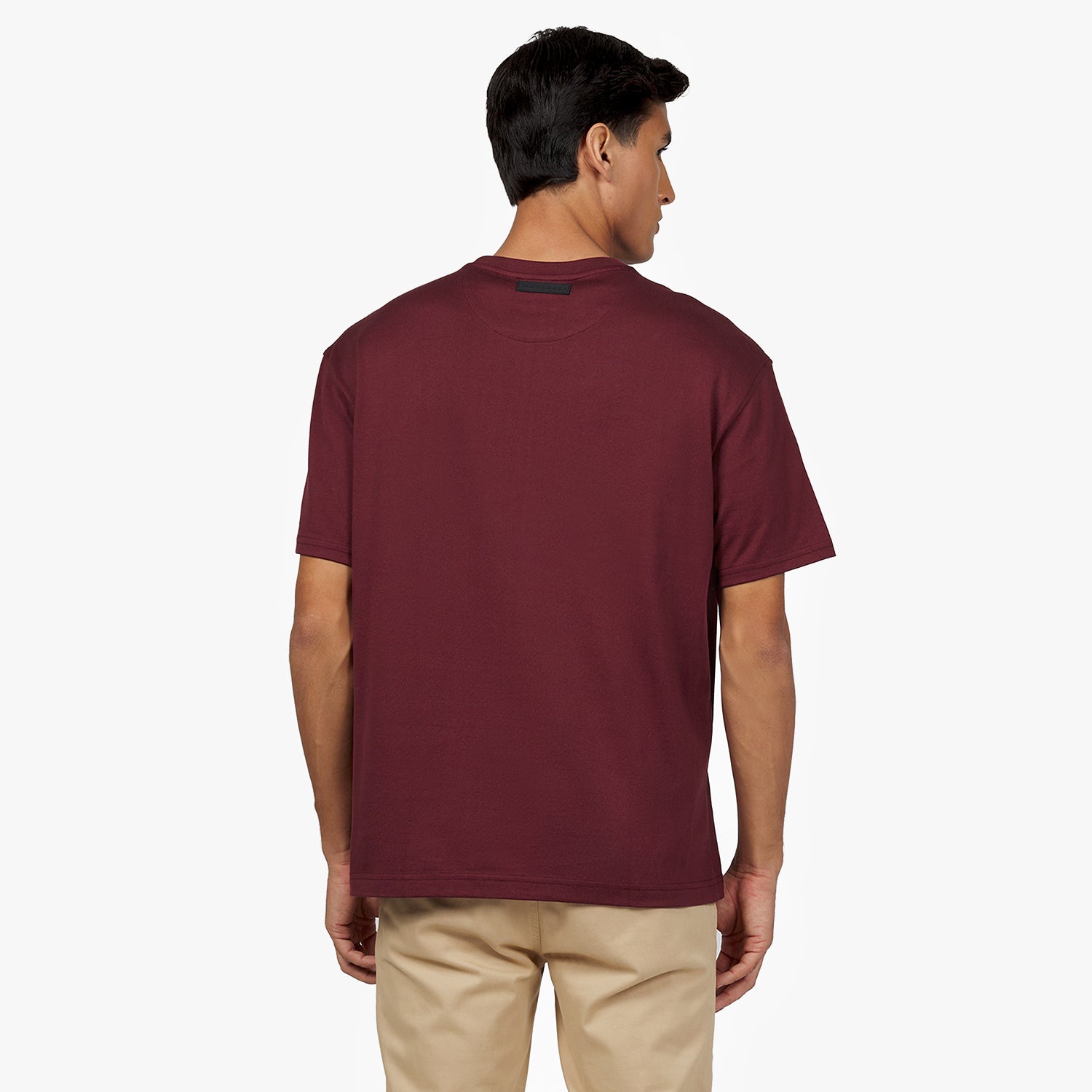 Back view of a male model wearing an oversized burgundy t-shirt. Features a minimalist design with a black rectangular 'JAMES BARK' label near the neckline. Paired with beige pants. White background