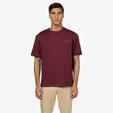 Male model wearing an oversized burgundy t-shirt with an embossed 'JAMES BARK ELEMENTS' logo on the chest. Features a ribbed crew neckline and short sleeves. Paired with beige pants. White background