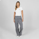 Full-length front view of a female model wearing a white crew neck t-shirt with a gold logo, wide-leg grey trousers, and white sneakers, standing against a light grey background