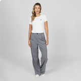 Full-length front view of a female model wearing a white crew neck t-shirt with a gold logo, wide-leg grey trousers, and white sneakers, standing against a light grey background