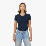 Front waist-up view of a female model wearing a navy blue crew neck t-shirt with a small gold embroidered logo on the left chest, paired with light wash jeans