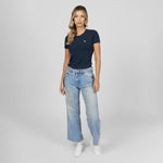 Full-length front view of a female model wearing a navy blue crew neck t-shirt with a gold logo, light wash jeans, and white sneakers, standing against a light grey background