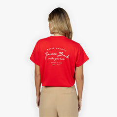 A young woman with wavy blonde hair is seen from the back wearing a red short-sleeved T-shirt featuring white printed text that reads 'PRIVÉ SOCIETY' at the top, followed by 'James Bark' in a script font, 'make your bark' underneath, and 'BASED IN USA EST. 2015' at the bottom. She pairs the T-shirt with high-waisted beige trousers and stands against a plain white background