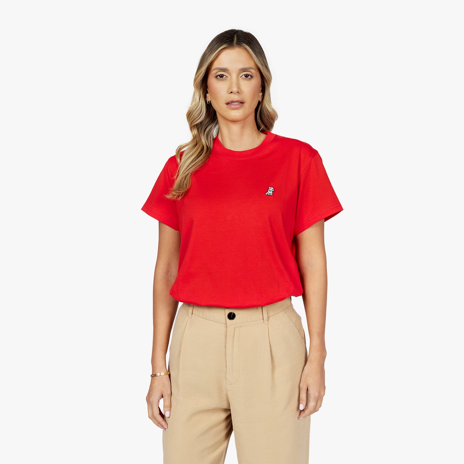 A young woman with wavy blonde hair wears a red short-sleeved T-shirt with a small embroidered white dog logo on the upper left chest area, tucked into high-waisted beige trousers. She stands against a plain white background with a neutral expression