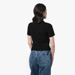 A female model with long, wavy black hair is shown from the back, standing against a plain white background. She is wearing a fitted, short-sleeved, black T-shirt with a classic round neckline. The back of the T-shirt is plain with no visible branding. She pairs it with medium-wash blue jeans that have a straight-leg fit and visible back pockets with contrast stitching. Her posture is relaxed, with her arms resting naturally at her sides while she slightly turns her head to the right.