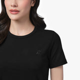 A close-up shot of a female model wearing a black, ribbed, short-sleeved T-shirt. The fabric has a fine ribbed texture, and the round neckline is neatly stitched. A small embroidered logo of a sitting dog, subtly blending with the fabric, is visible on the left side of the chest. Part of the model’s face is shown, revealing her lips, chin, and dark wavy hair resting over her shoulder.