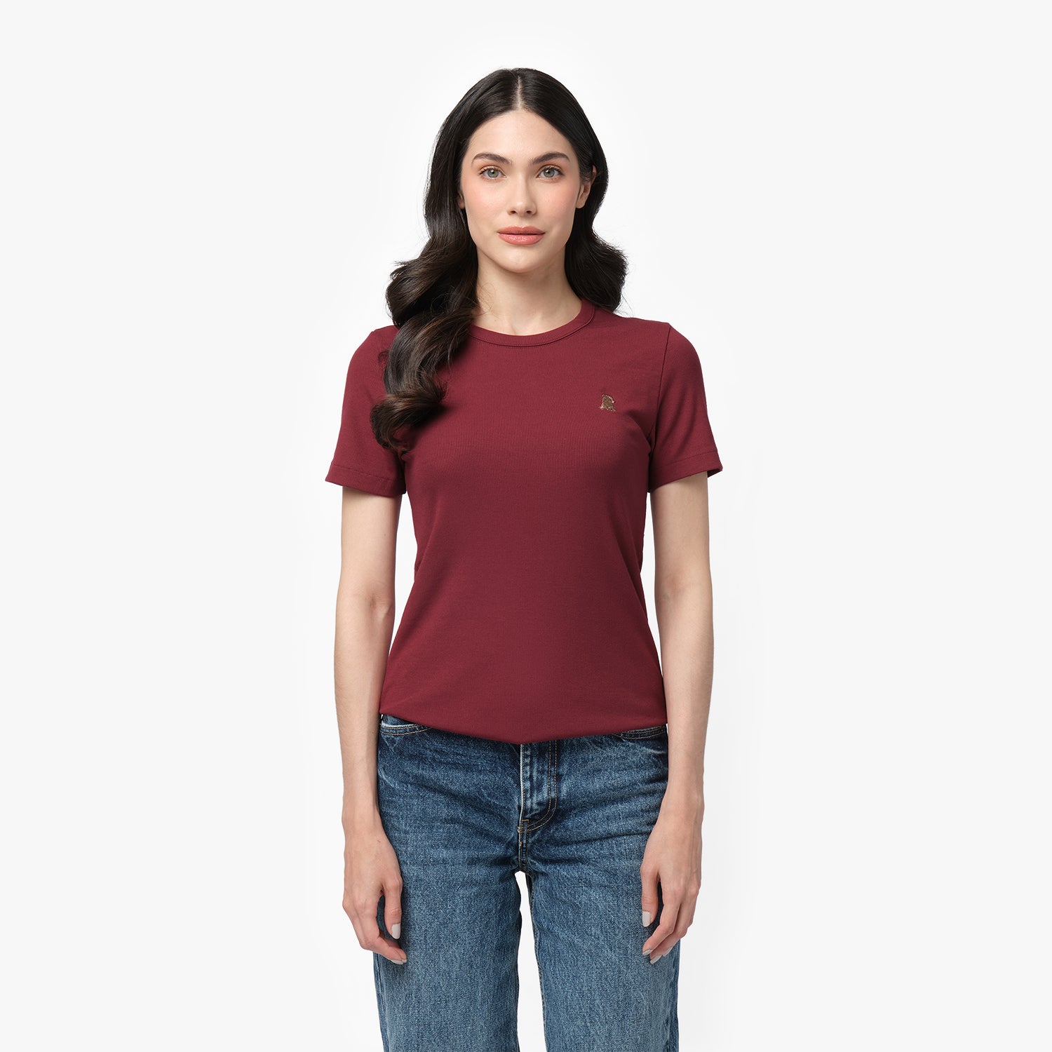 A female model with long, wavy black hair stands against a plain white background. She is wearing a fitted, short-sleeved, burgundy T-shirt with a small embroidered logo of a sitting dog on the left chest area in a slightly darker shade. The T-shirt has a classic round neckline and a smooth fabric texture. She pairs it with high-waisted, medium-wash blue jeans in a straight-leg fit. Her expression is neutral with a hint of a smile, and her posture is relaxed, with her arms resting naturally at her sides.