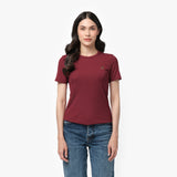 A female model with long, wavy black hair stands against a plain white background. She is wearing a fitted, short-sleeved, burgundy T-shirt with a small embroidered logo of a sitting dog on the left chest area in a slightly darker shade. The T-shirt has a classic round neckline and a smooth fabric texture. She pairs it with high-waisted, medium-wash blue jeans in a straight-leg fit. Her expression is neutral with a hint of a smile, and her posture is relaxed, with her arms resting naturally at her sides.