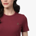 A close-up shot of a female model wearing a burgundy, ribbed, short-sleeved T-shirt. The fabric has a fine ribbed texture, and the round neckline is neatly stitched. A small embroidered logo of a sitting dog, slightly darker than the fabric, is visible on the left side of the chest. Part of the model’s face is shown, revealing her lips, chin, and dark wavy hair cascading over her shoulder.