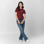 A female model with long, wavy black hair poses against a plain light gray background. She is wearing a fitted, short-sleeved, burgundy T-shirt with a small embroidered logo of a sitting dog on the left chest area. The T-shirt is paired with high-waisted, medium-wash blue jeans in a relaxed, straight-leg fit. She completes the outfit with white sneakers. Her pose is casual and elegant, with her legs crossed at the ankles and her hands gently resting in front of her while she looks slightly to the side with 