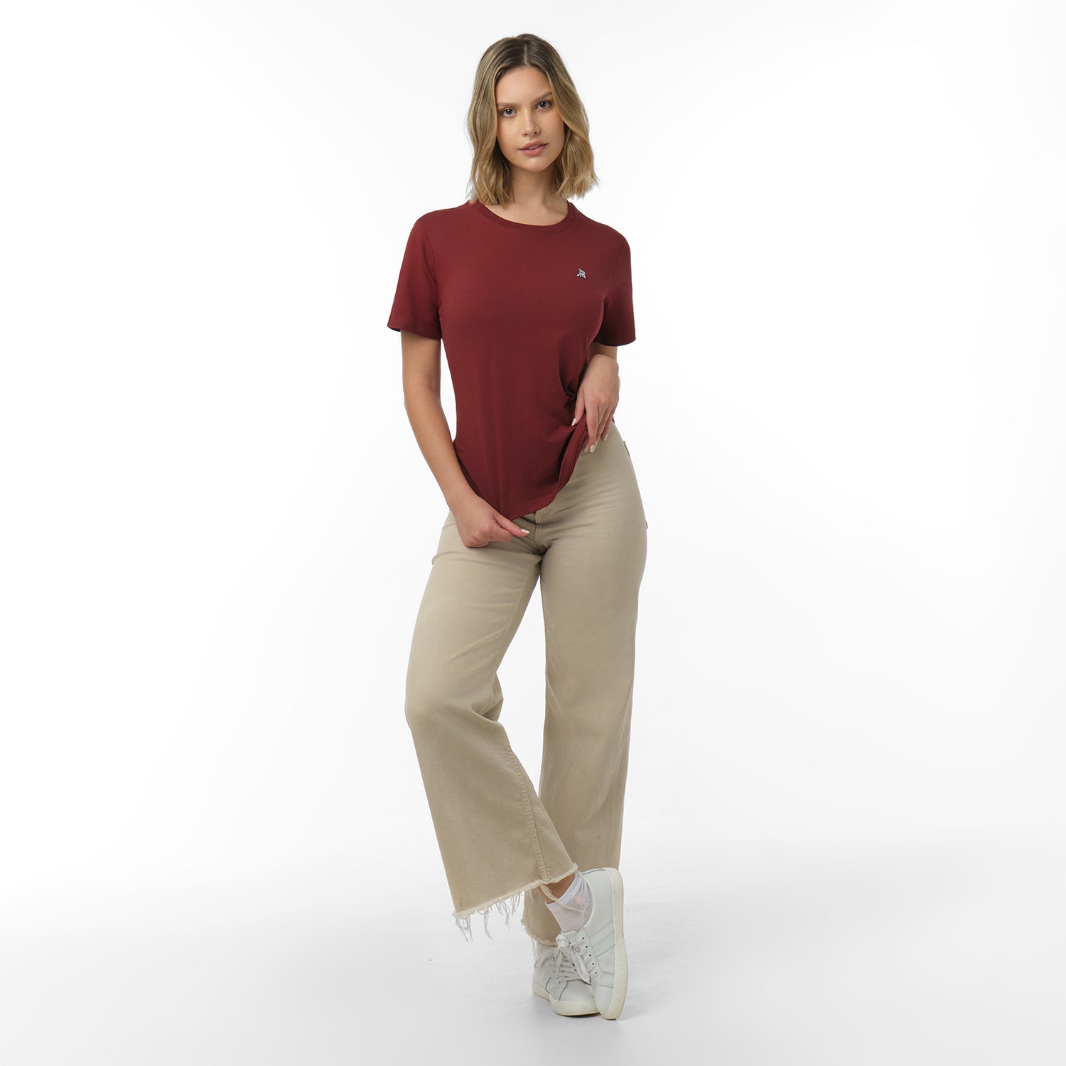 Woman standing full length, posing in a maroon t-shirt with small white logo, beige wide-leg pants with frayed hem, white socks, and white sneakers