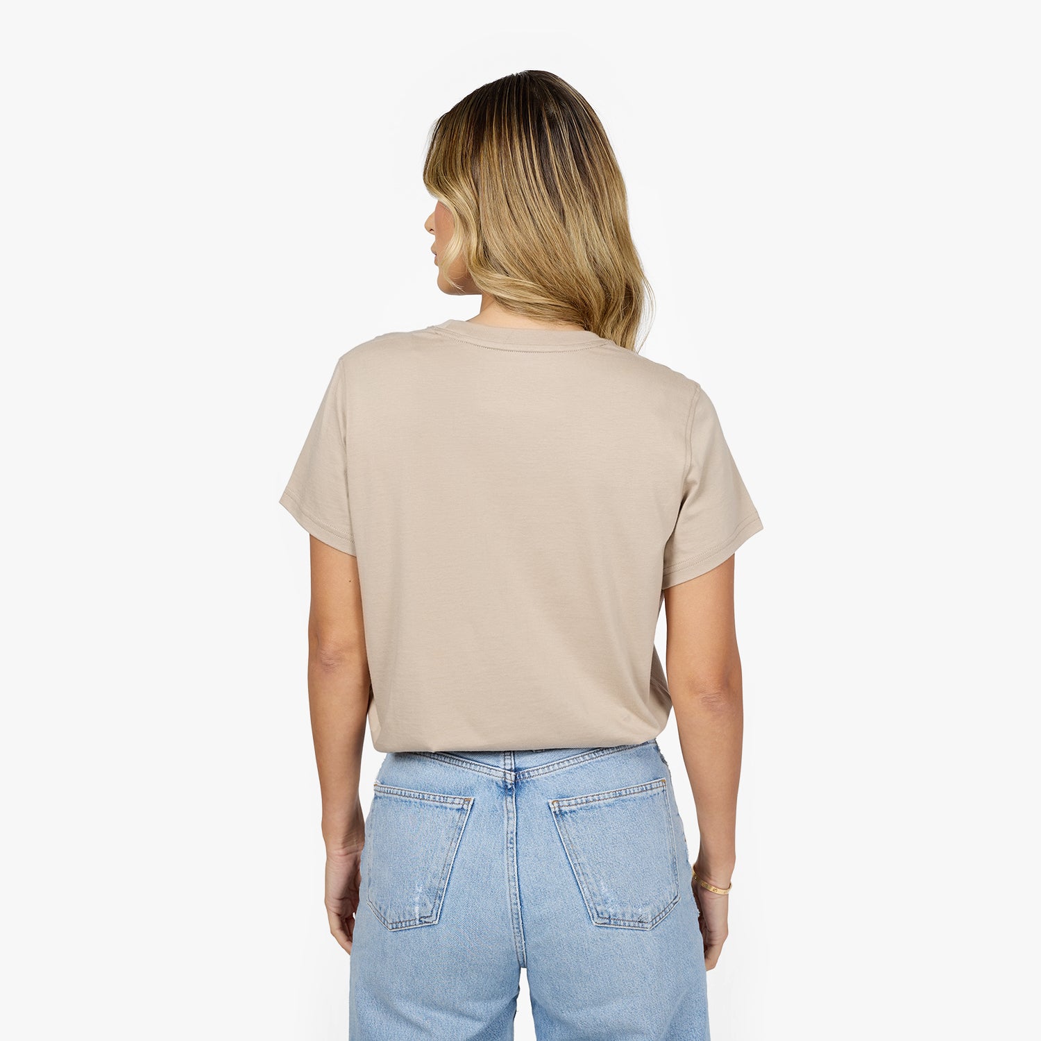 A woman with long, wavy blonde hair is shown from the back, wearing a beige oversized t-shirt and high-waisted, light-wash blue jeans. The t-shirt has a relaxed fit with short sleeves and no visible 