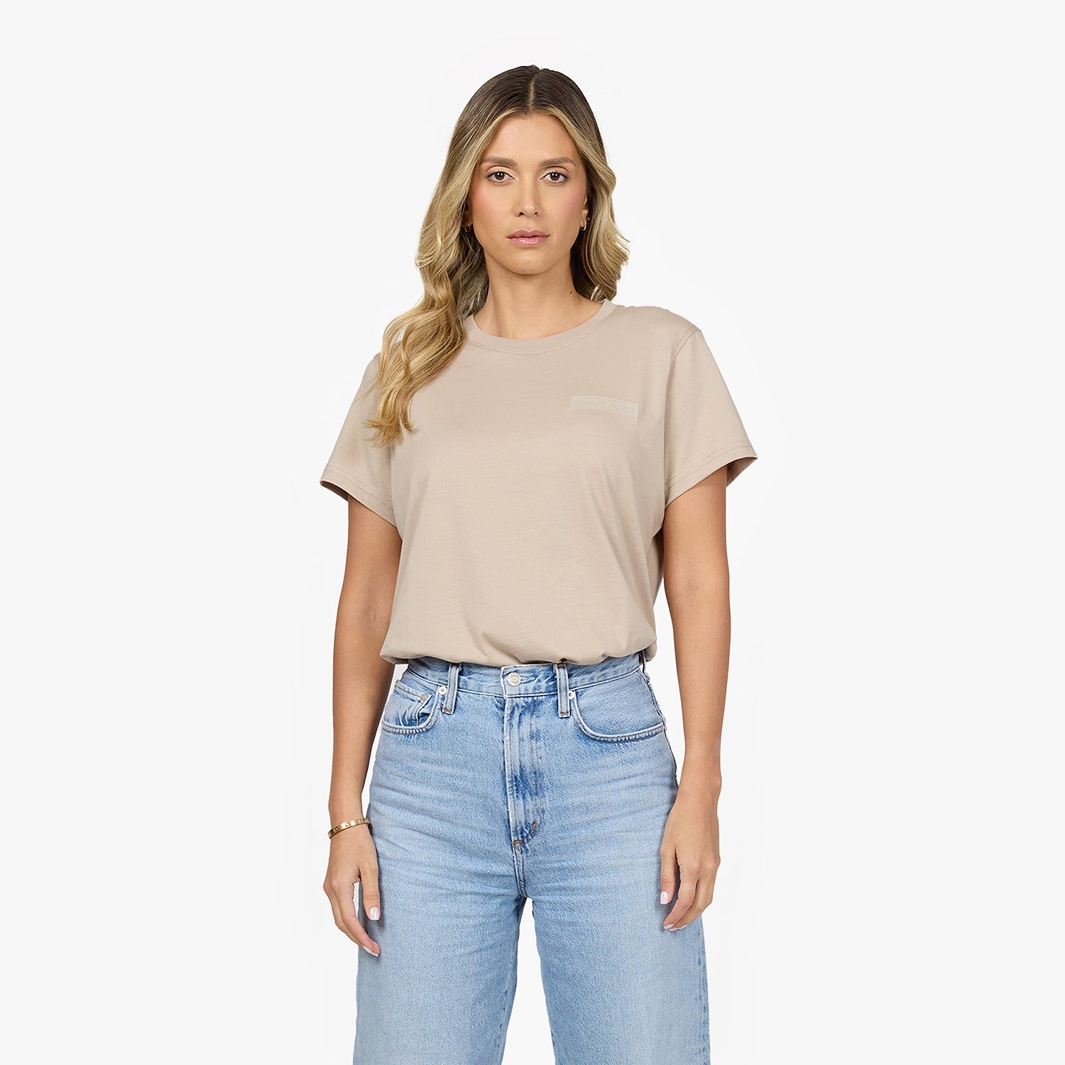 A woman with long, wavy blonde hair wears a beige oversized t-shirt tucked into high-waisted, light-wash blue jeans. The t-shirt features a subtle embossed logo on the left chest. She has a neutral expression, gold hoop earrings, and a delicate gold