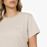 A close-up of a woman wearing a beige t-shirt with a minimalist embossed logo on the left chest area reading "PRIVÉ SOCIETY JAMES BARK EST. 2015." The fabric appears soft, and the short sleeves have a clean hem. The woman has wavy blonde hair and wears subtle gold hoop earrings. The background is plain white.