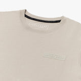 A close-up of a beige T-shirt, highlighting the tonal rubberized logo patch on the upper left chest that reads 'PRIVÉ SOCIETY JAMES BARK EST. 2015.' The neckline features a black inner lining with repeated 'JAMES BARK' branding, and the inside label displays 'JAMES BARK MAKE YOUR BARK' along with material details