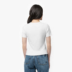 A female model with long, wavy black hair is shown from the back, standing against a plain white background. She is wearing a fitted, short-sleeved, white T-shirt with a classic round neckline. The back of the T-shirt is plain with no visible branding or embellishments. She pairs it with high-waisted, medium-wash blue jeans that have a straight-leg fit and visible back pockets with contrast stitching. Her posture is relaxed, with her arms resting naturally at her sides while she slightly turns her head to t