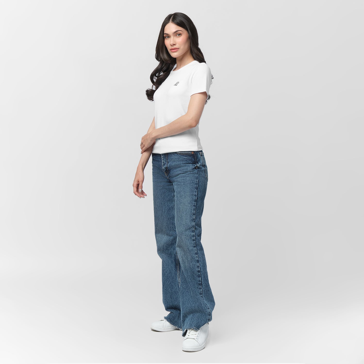 A female model with long, wavy black hair poses against a plain light gray background. She is wearing a fitted, short-sleeved, white T-shirt with a small embroidered logo of a sitting dog on the left chest area in black. The T-shirt is paired with high-waisted, medium-wash blue jeans in a relaxed, straight-leg fit. She completes the outfit with white sneakers. Her pose is casual, standing at a slight angle with her left arm gently touching her right forearm while looking slightly to the side with a neutral 