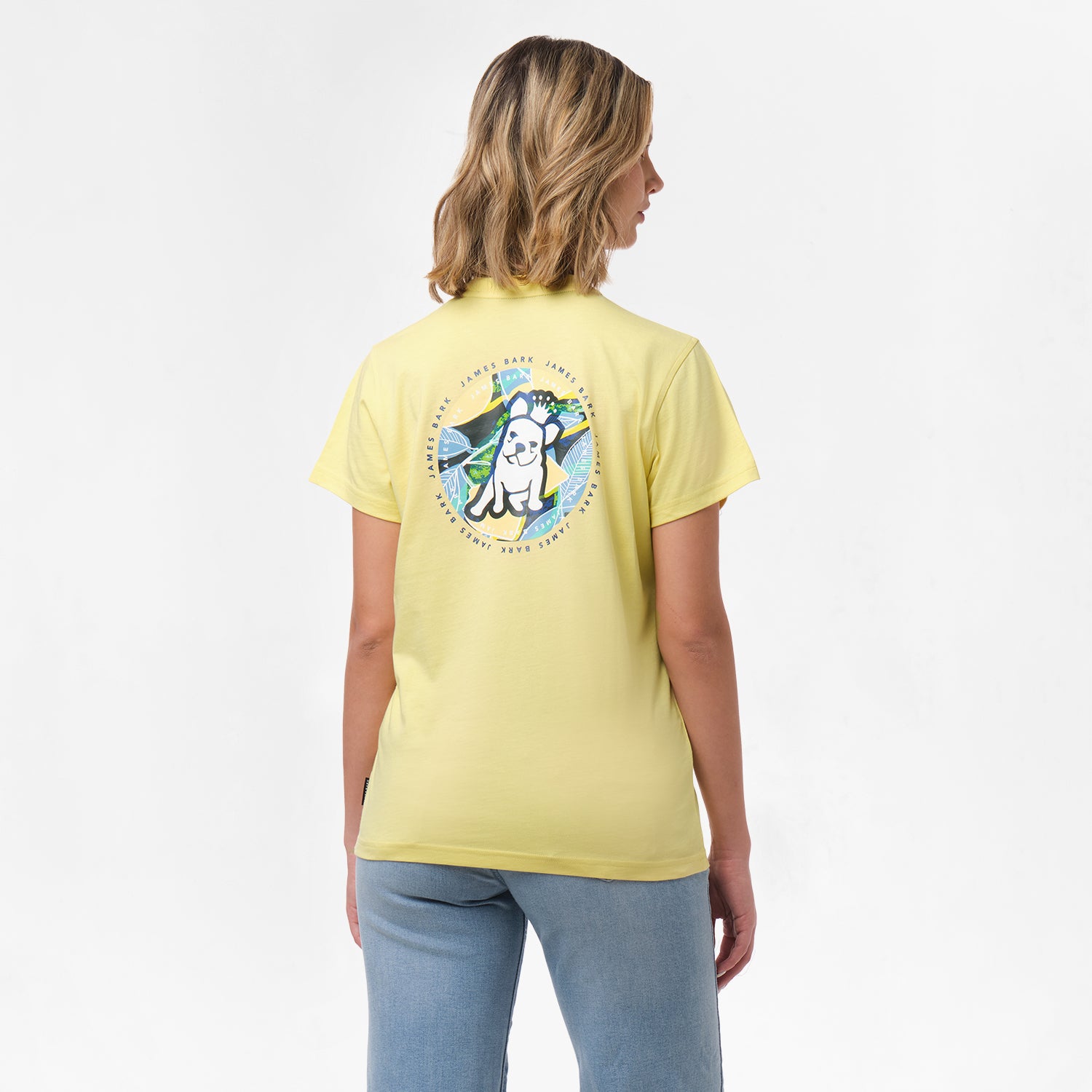 A woman is shown from behind, wearing a yellow T-shirt with a large circular graphic on the back. The design features a dog sitting in front of a tropical background with the text "James Bark Privé Society" around the circle. She pairs the shirt with light blue jeans.