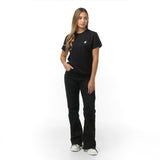 A woman stands against a plain white background, wearing a black T-shirt with a small embroidered dog logo on the chest. She pairs the shirt with black pants and white sneakers. She has long, wavy hair and stands with one hand in her pocket, looking slightly to the side with a neutral expression.