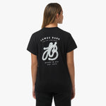 A woman is shown from behind, wearing a black T-shirt with a large white graphic on the back. The design features the text "James Bark Privé Society," with a rope illustration forming the initials "JB" and the words "Based in USA, Est. 2015" below. She pairs the shirt with black pants.