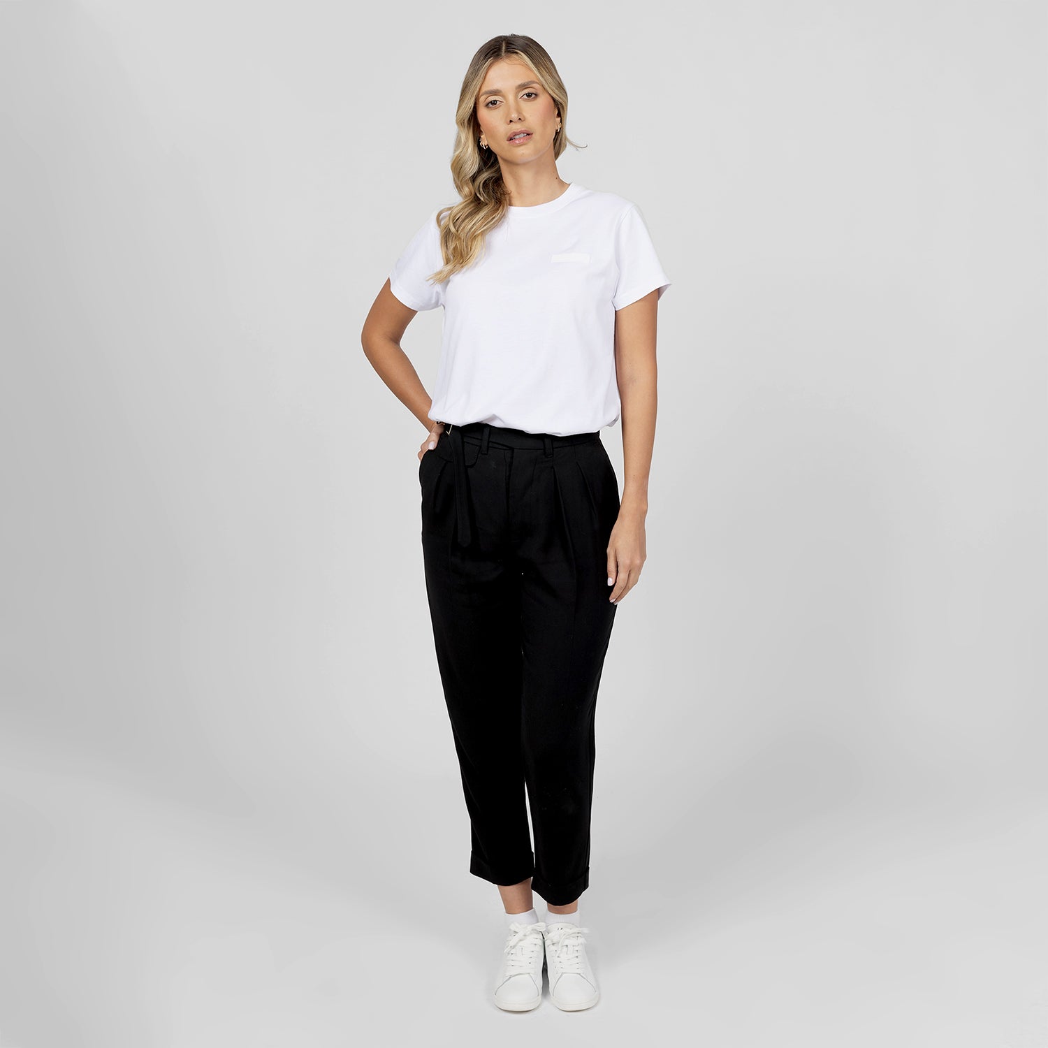 A woman with wavy blonde hair poses in a minimalistic outfit consisting of a plain white t-shirt with a subtle embossed logo and tailored black trousers with a belted waist. She completes the look with white sneakers, giving a casual yet sophisticated vibe. Her relaxed stance and neutral expression complement the clean and modern aesthetic. The background is a solid light gray, enhancing the simplicity of the outfit.