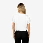 A woman with blonde wavy hair is seen from the back wearing a plain white t-shirt with a relaxed fit, tucked into high-waisted black trousers with buttoned back pockets. She accessorizes with a gold bracelet. The plain white background emphasizes the minimalist and classic style of the outfit