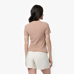 A female model with long, wavy black hair is shown from the back, standing against a plain white background. She is wearing a short-sleeved, beige T-shirt with a simple, smooth design and no visible branding on the back. The T-shirt is paired with off-white, high-waisted shorts with a slightly loose fit. Her posture is relaxed, with her arms naturally resting at her sides while she looks slightly to the side.