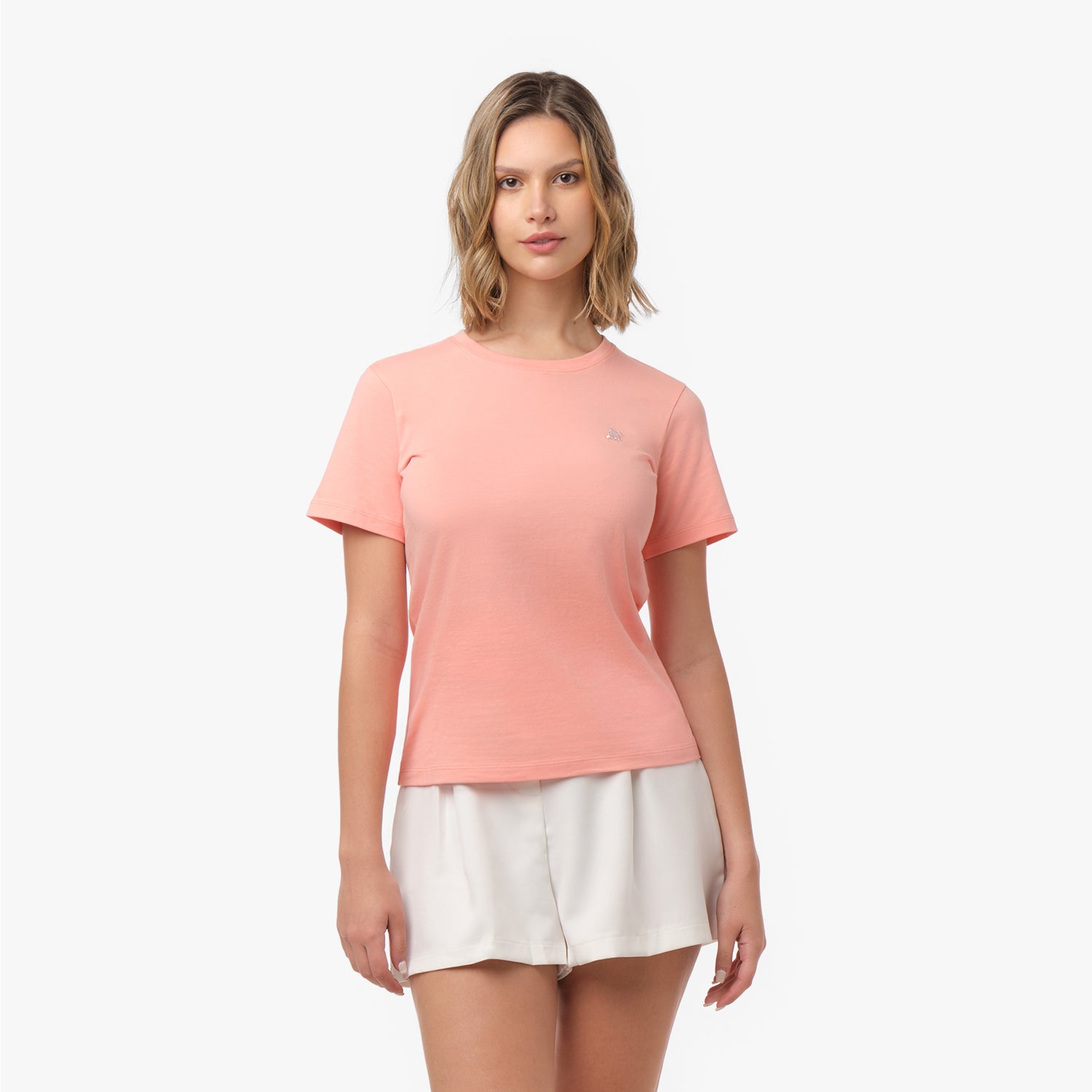 Woman facing forward wearing a peach-colored t-shirt with small tone-on-tone embroidered dog logo on left chest and white shorts