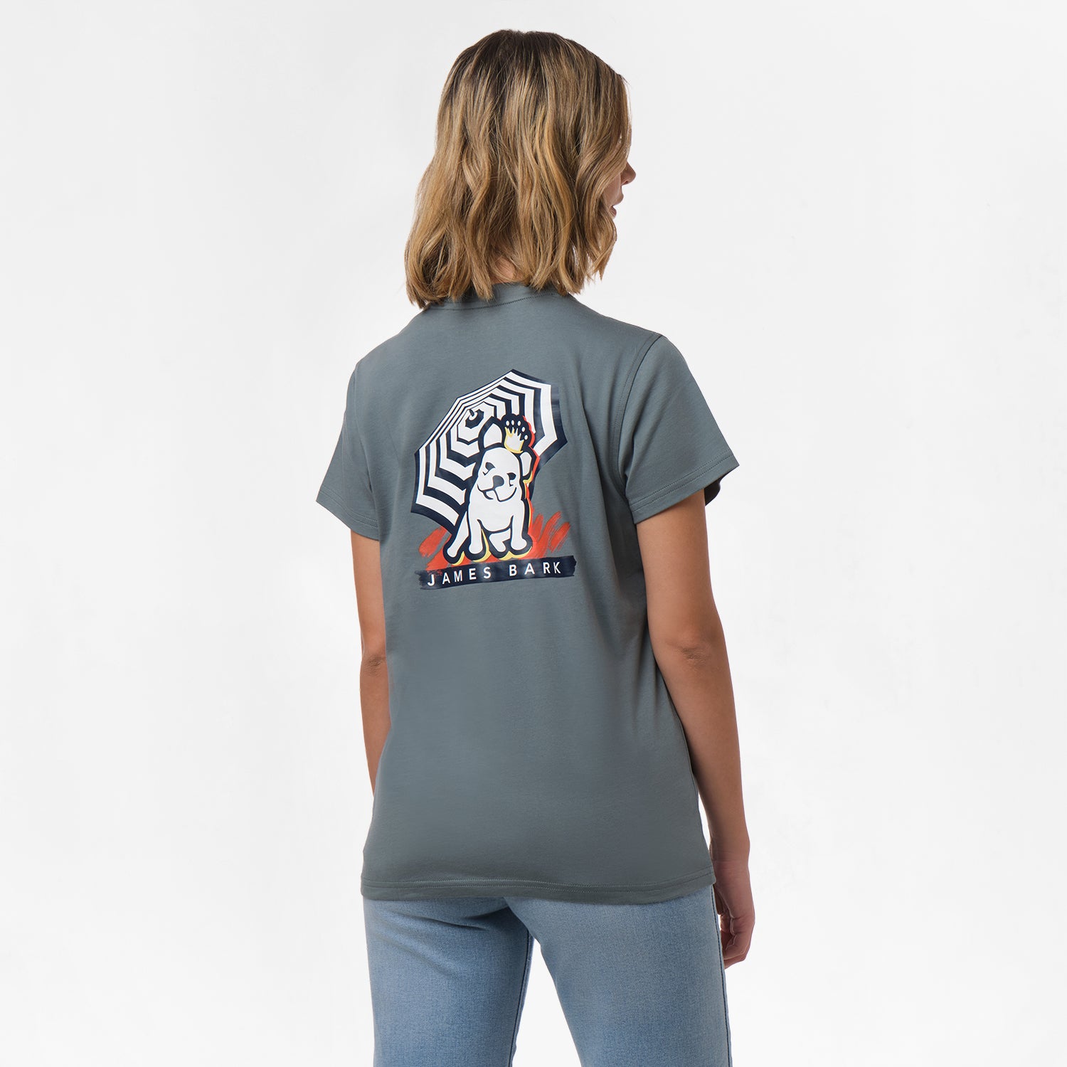 A woman is shown from behind, wearing a gray T-shirt with a large graphic on the back. The graphic features a dog sitting under a striped umbrella with the text "James Bark" below. She pairs the shirt with light blue jeans.

