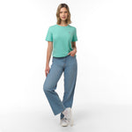 Woman standing full length, posing with one leg crossed and hand in pocket, wearing a mint green t-shirt with white logo, blue wide-leg jeans with frayed hem, white socks, and white sneakers