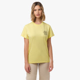 A woman stands facing forward, wearing a yellow T-shirt with black text on the chest that includes phrases like "Dolce Vita," "Amore," and "Amo Stupendo." She pairs the shirt with beige pants and has a neutral expression.